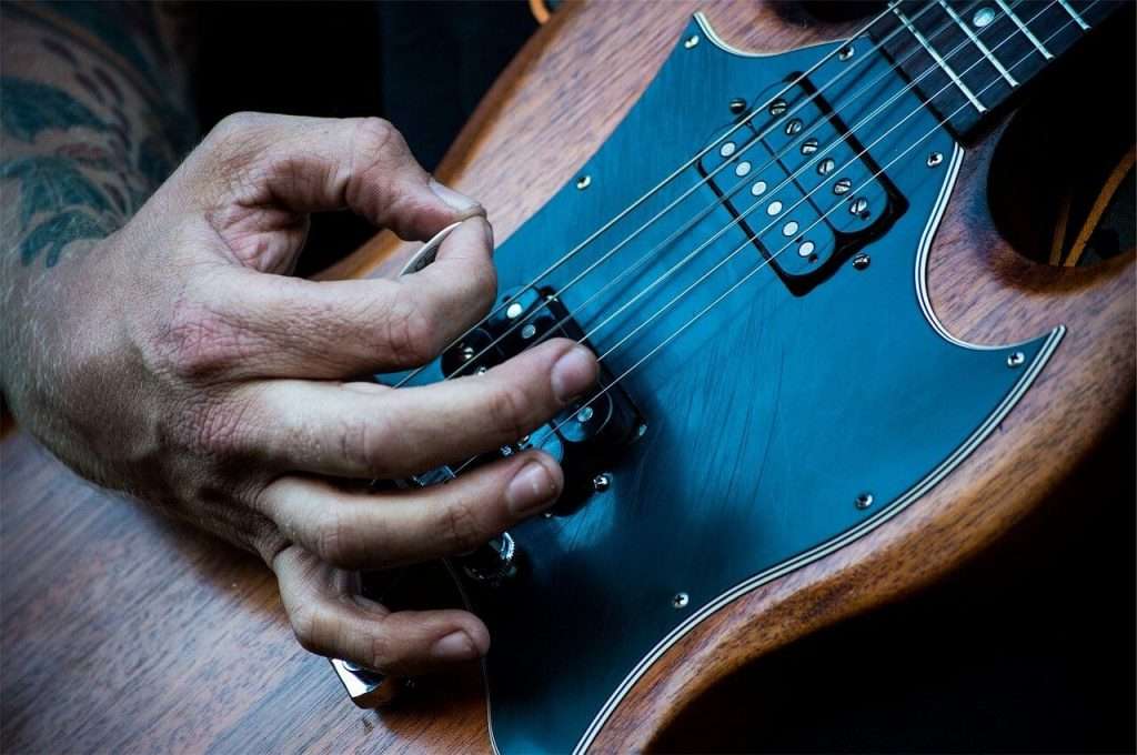 Person playing the electric guitar: zoomed on the hand holding the pick near the strings, the pickups and the pickguard.